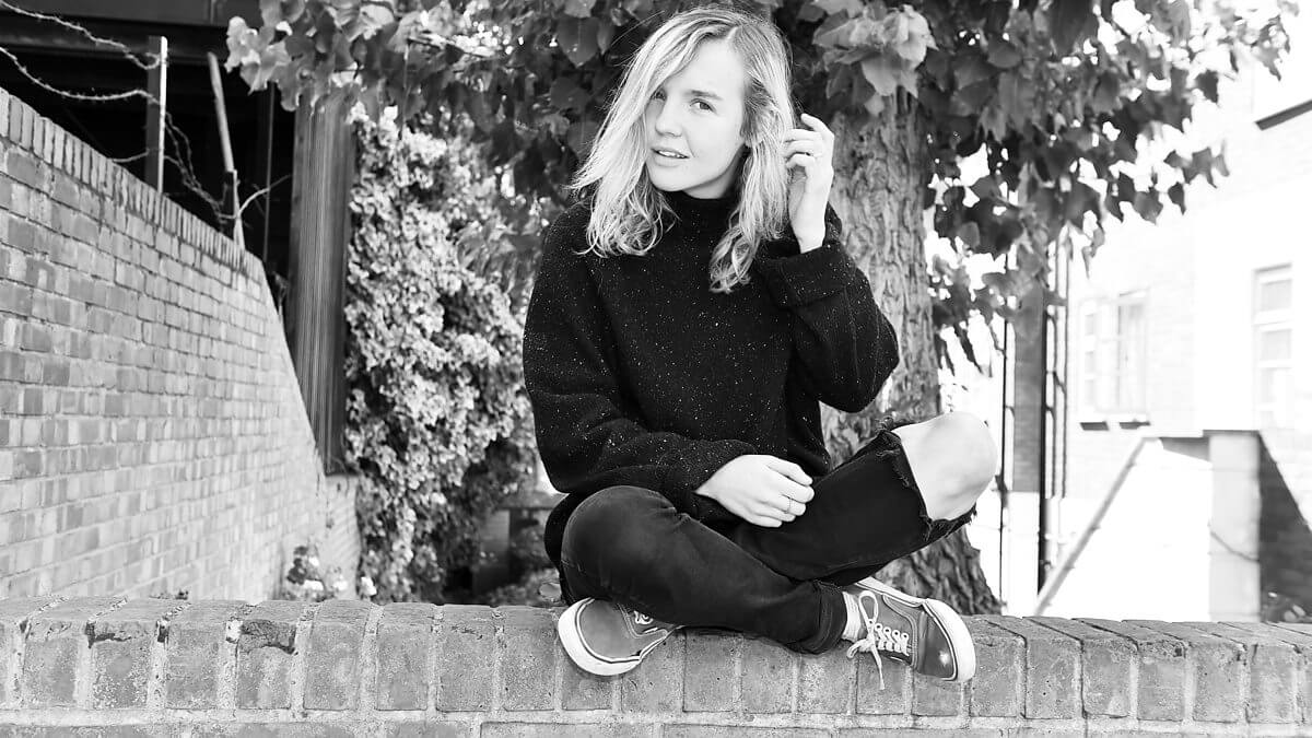 Potret hitam putih The Japanese House dengan sweater dan jeans sambil duduk di atas tembok batu bata